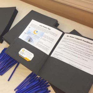 A black folder with a questionnaire, a colorful rack card and a business card lays on a table with pens for a seminar.
