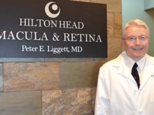 Dr. Peter Liggett stands wearing a white coat in front of his practice's sign in the waiting room