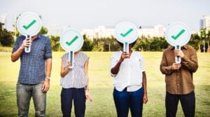 Four people stand in a park near a city skyline holding signs with checkmarks over their faces.