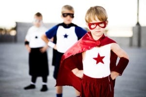 A little boy with a red cape and mask stands in the forefront of two other little boys dressed as super heroes