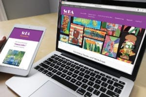 An open laptop displays a colorful website for a non-profit art association and a tablet held by a hand shows an blast